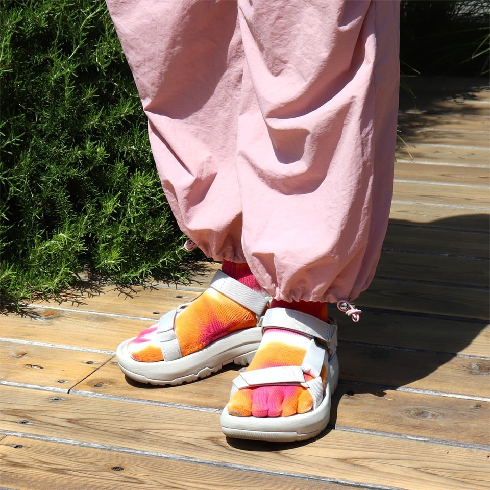 Colorful sandals worn on a wooden path with pink pants and greenery.