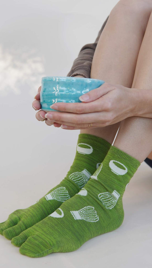 Person wearing green socks with bamboo whisk patterns, holding a blue matcha bowl.