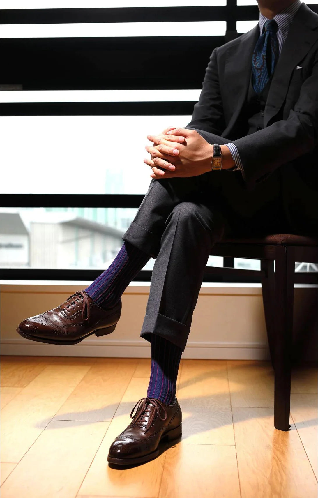 A man wearing brown leather shoes and a gray suit crosses his legs over a pair of Il Regalo Shadow Ribbed Mid-Calf Socks in Charcoal Grey