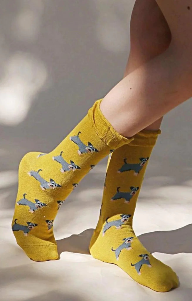 Side view of a woman's leg wearing socks with a yellowish mustard-colored fabric of a product called Animal Rescue Pairs SCHNAUZER Dog Socks from the Tabbisocks brand with an illustration of a miniature schnauzer throughout