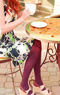 A woman relaxes in a cafe wearing a pair of Tabbisocks 23 Colors Opaque Zokki Tights in Merlot with a green and navy blue patterned dress and beige sandals